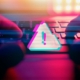 A stylized image showing hands typing on a keyboard with a glowing warning icon overlay, representing cyber threat activity. The visual includes glitch effects and intense lighting, symbolizing digital risk. Ideal for content related to unified threat management in Dallas.